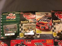 Photo showing packaged diecast NASCAR Sprint Cup Series cars with collector cards and display stands, along with NFL Racing collector car and card set.