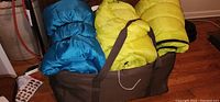 Blue and yellow sleeping bags rolled and secured with straps inside a large brown tote on wooden floor.