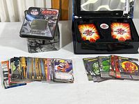 Photo shows Bakugan Battle Brawlers and Naruto trading cards alongside their themed storage containers including a Naruto metal tin box and a black card case with Bakugan logo. Cards include ability and special effect cards typical to the Bakugan game.