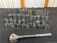 Full set of various size sockets arranged in rows with one chrome ratchet wrench in front on black fabric.