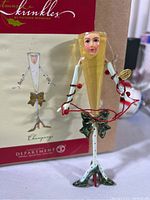 Close-up of Department 56 Krinkles champagne glass ornament showing whimsical female figure with cream dress, red berries, and green holly bow detail. Original packaging visible in background.