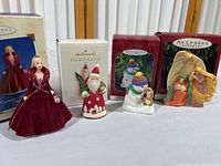Four Hallmark Keepsake Christmas ornaments including a Barbie doll in a velvet dress, a Santa figure, a snowman with a dog, and an angel nativity scene with original boxes