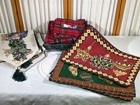 Photo showing a collection of holiday-themed placemats, napkins, and table runners on a white surface, displaying woven and embroidered Christmas ornament designs and a cream table runner with tassels.
