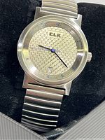 Close-up of watch dial showing silver textured patterned face, CLK logo, blue second hand, black hour and minute hands, and date window at 6 o'clock.