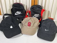 Full view showing all items: two Nike pouches and three hats with Apple, Stanford, and Nike logos on a white surface.