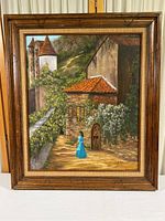 Framed oil painting showing entire artwork including wood frame and burlap mat, depicts a woman in blue walking beside old stone buildings with greenery.