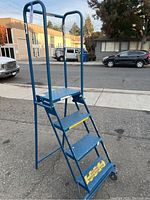 Full view of blue metal rolling ladder showing four steps, tubular handrail, wheels at base
