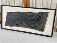 Frontal view of the black mink carved in slate mounted on white painted cedar plank with a slim dark border frame.