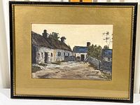 Framed watercolor painting showing an older style gold-toned frame and tan mat around a farm scene with stone buildings and trees, signature visible lower left.