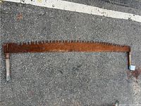 Full view of the large vintage two-man crosscut saw laid flat on pavement showing the entire rusted saw blade and wooden handles.