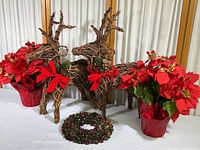 Two woven wood deer, two faux potted poinsettia plants wrapped in red foil, and a small Christmas wreath shown on a white surface in front of a curtain.