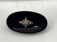 Front view of the oval black onyx brooch with central pearl and gold decoration on white fabric.