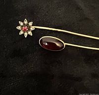 Close-up view of the two stick pins on a dark background, showing the floral ruby pearl pin and the oval red resin pin.
