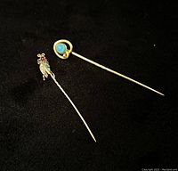 Two yellow gold stick pins shown on black background, one with a round blue stone in circular setting, the other insect shaped with multi-colored stone inlays.