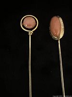 Two vintage 14 kt yellow gold stick pins with coral stones displayed on black background. One pin has round coral stone in a simple gold bezel, the other an oval coral stone in an engraved gold bezel.