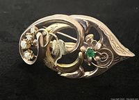 Front view on black background showing open scrollwork, emerald center stone, pearl cluster and red accent