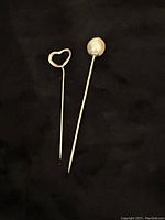 Two stick pins placed on black fabric backdrop, one with hollow heart top and one with pearl top
