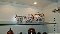 Glass shelf displays three-piece Royal Albert style set: teapot, sugar bowl with lid, and creamer with matching floral pattern and gold trim under shelf lighting.