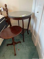 Two round wooden tables in dark brown finish placed next to a bed. The smaller table has a tilt top in open position, resting on three curved legs. The larger round table has a glass top and curved legs with a scalloped lower shelf.