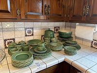 Overview of green majolica plates, cups, mugs, serving pieces and cookie jar on tiled counter