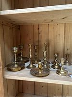 Wide view showing all 10 assorted brass candle holders on wooden shelf, various styles and heights.