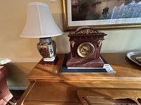 Wider angle photo showing both marble clock and floral ceramic lamp on a polished wooden furniture surface in a room setting.