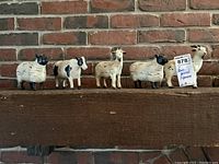 All five farm animal figurines arranged in a row on a wooden surface, showing their relative sizes and styles against a brick wall backdrop.