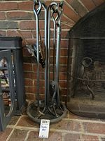 Full view of the wrought iron fireplace tool set standing on a brick hearth, showing the stand and all hanging tools.