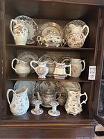 Full set of ceramic items displayed in a wooden cabinet including pitchers, plates, tureen, and candle sticks all featuring brown floral patterns.