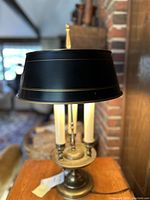 Side and angled view of brass-like table lamp with black shade, showing three white faux candle-style lights and circular base.