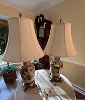 Pair of floral porcelain table lamps on a table, showing the full lamp with beige fabric shades and floral decorated bases with gold trim.