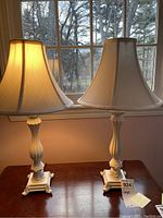 Two off white table lamps on wooden table by window, one lamp lit showing warm glow, both with fabric shades and ornate bases.