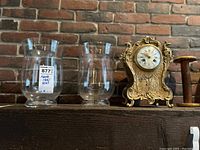 Full shot of two clear glass candle holders and gold ornate mantel clock on wooden shelf with brick background.