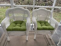 Wide front view of both white plastic wicker chairs with green cushions placed side by side on a porch.