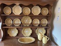 Wooden hutch displaying mustard-brown cabbage leaf plates, cream scalloped plates, large leaf plates, oval platter, and two pitchers