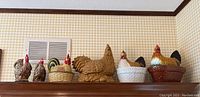 Seven ceramic rooster pieces including two casserole dishes and five figurines, displayed on a shelf against a checkered wallpaper background.