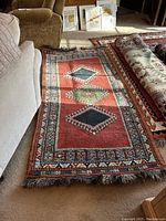 Photo of the full length of the worn Oriental style rug against carpeted floor showing central diamond patterns and fringed edges.