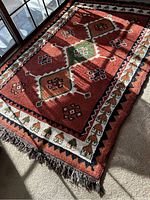 Full view of oriental style rug laid flat showing central medallion and surrounding geometric patterns in red, green, brown, white, black colors, with fringed edges.