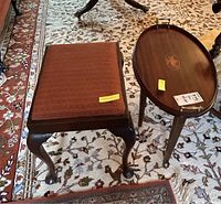 Wooden stool with upholstered rust-colored seat and claw-foot legs next to oval wooden side table with inlaid design on top