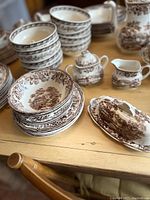 View of stacked bowls, plates, covered butter dish, sugar bowl, and creamer all with brown transferware pattern on table.