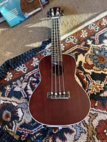 Front view of mahogany finish ukulele with four nylon strings and blue/black patterned strap, on a patterned rug.