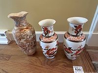 Photo of three vases together on wooden floor: two similar white vases with floral patterns and one larger beige detailed vase