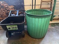 Photo showing both the black Smart Cart 450 wheelbarrow and green plastic yard waste barrel side by side indoors with a stack of firewood in background.