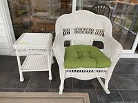 White wicker rocking chair with green cushion next to white wicker side table on patio floor with tile.