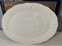 Top-down view showing the white ceramic platter with raised turkey motif and leaf-patterned rim against its original box.