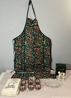 Photo showing the Christmas apron hanging on the wall, two white embroidered hand towels, six holiday glasses, and the boxed Snowbabies figurine on a table covered with white cloth.
