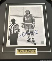 Front view of framed autographed photo showing Dennis Maruk in California Golden Seals uniform on the ice next to referee, with signature.