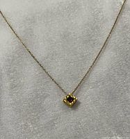 Close-up image showing pendant detail: square gold-tone pendant with textured edges and faceted ruby-like red stone center on a delicate chain.