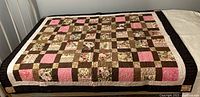 Full view of handmade patchwork quilt laid flat showing checkerboard pattern with floral, solid pink, solid brown, and tan patches and a dark brown border.