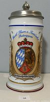 Front view of the beer stein showing detailed color crest with crown and shield on ceramic body with blue and gold accents.